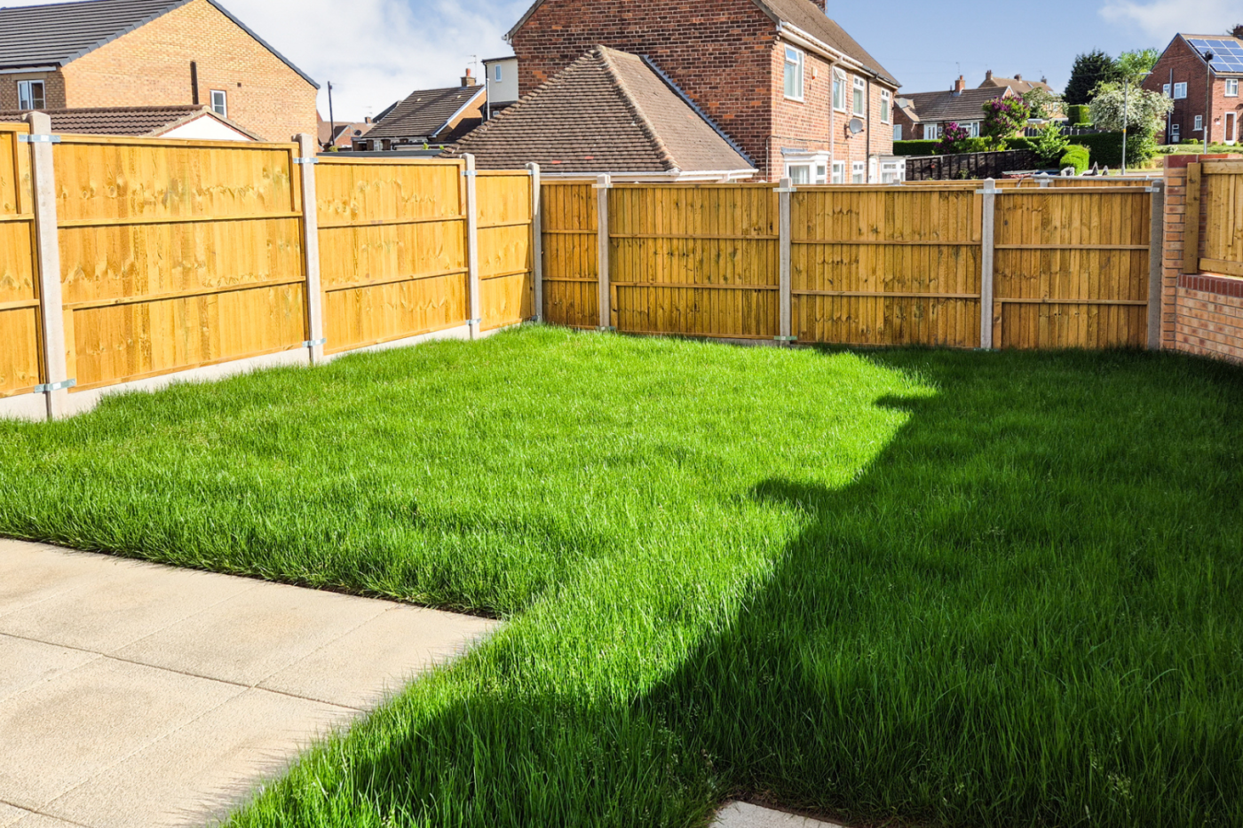 The garden showing green grass, patio slabs, the garden fence and a fence sitting on top of a small brick wall.
