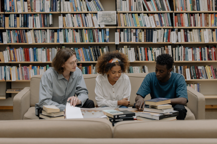 3 students sitting on a sofa in a library looking at books together