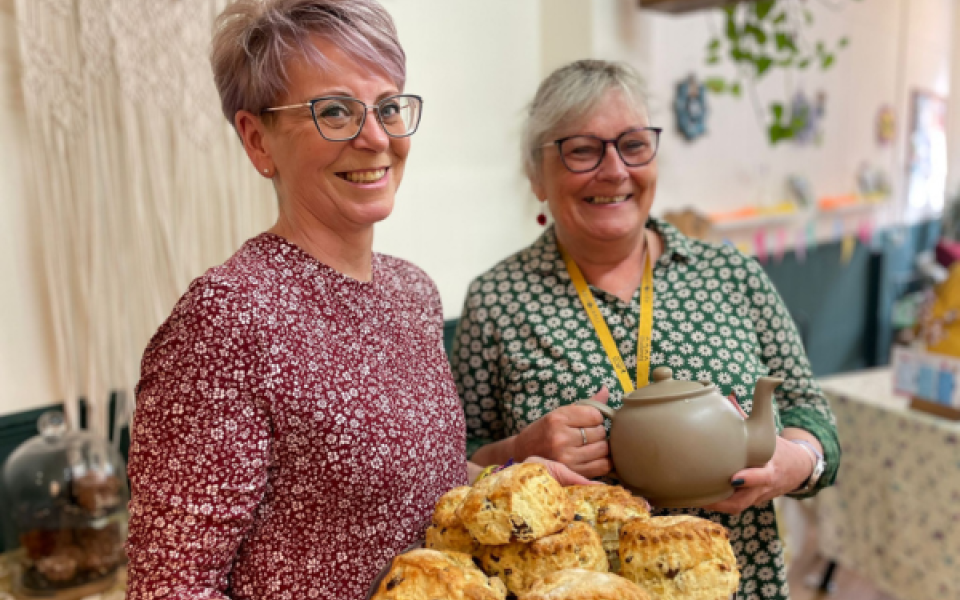 Two ladies  smiling holding a teapot and scones