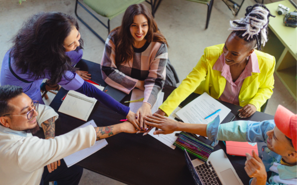 A diverse group of people sat in a circle with their hands on top of one another in the middle.